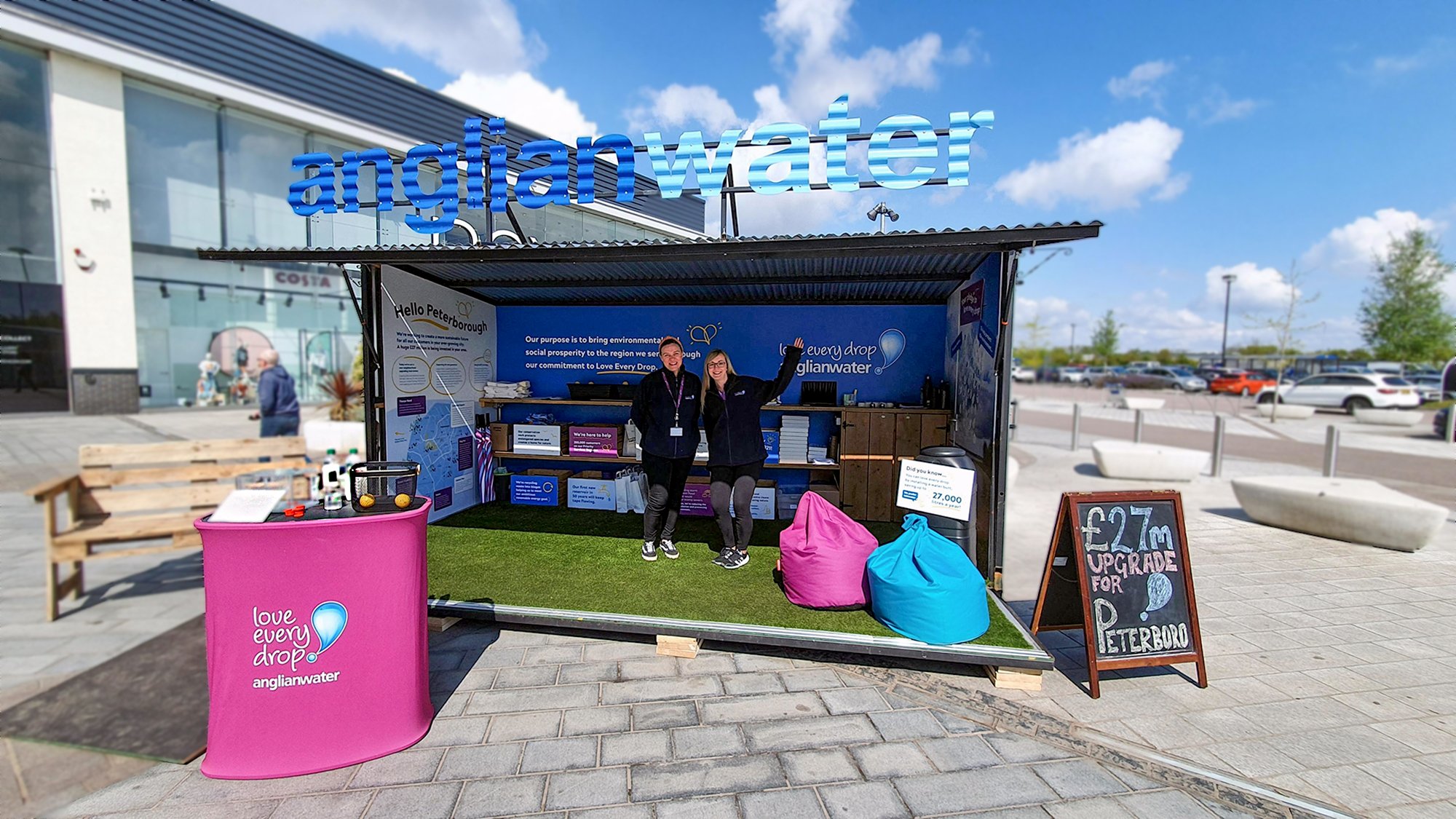 A promotional booth for Anglian Water with a large blue sign on top. Two people are standing inside the booth, smiling and waving. The booth has informational posters, gift bags, and a pink and a blue bean bag chair outside. A chalkboard sign advertises a £27 upgrade for Peterborough. The booth is set up outdoors on a paved area with parking and a building in the background.