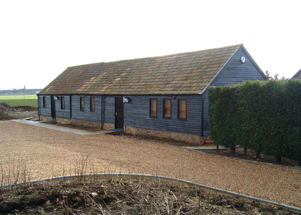 A small, single-story house with a dark blue wooden exterior and a pitched roof, surrounded by a gravel driveway and some greenery on one side.