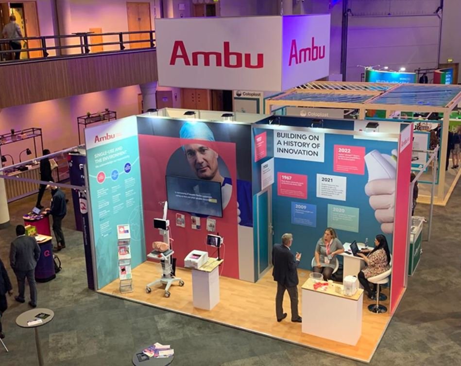 Trade show booth for Ambu, with large banners displaying the company's name and focus on innovation; features medical equipment and informational displays, with people engaging in conversations and viewing products.