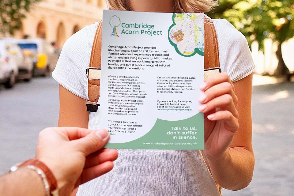Person holding a flyer for the Cambridge Acorn Project outdoors, with a background of trees and parked cars.