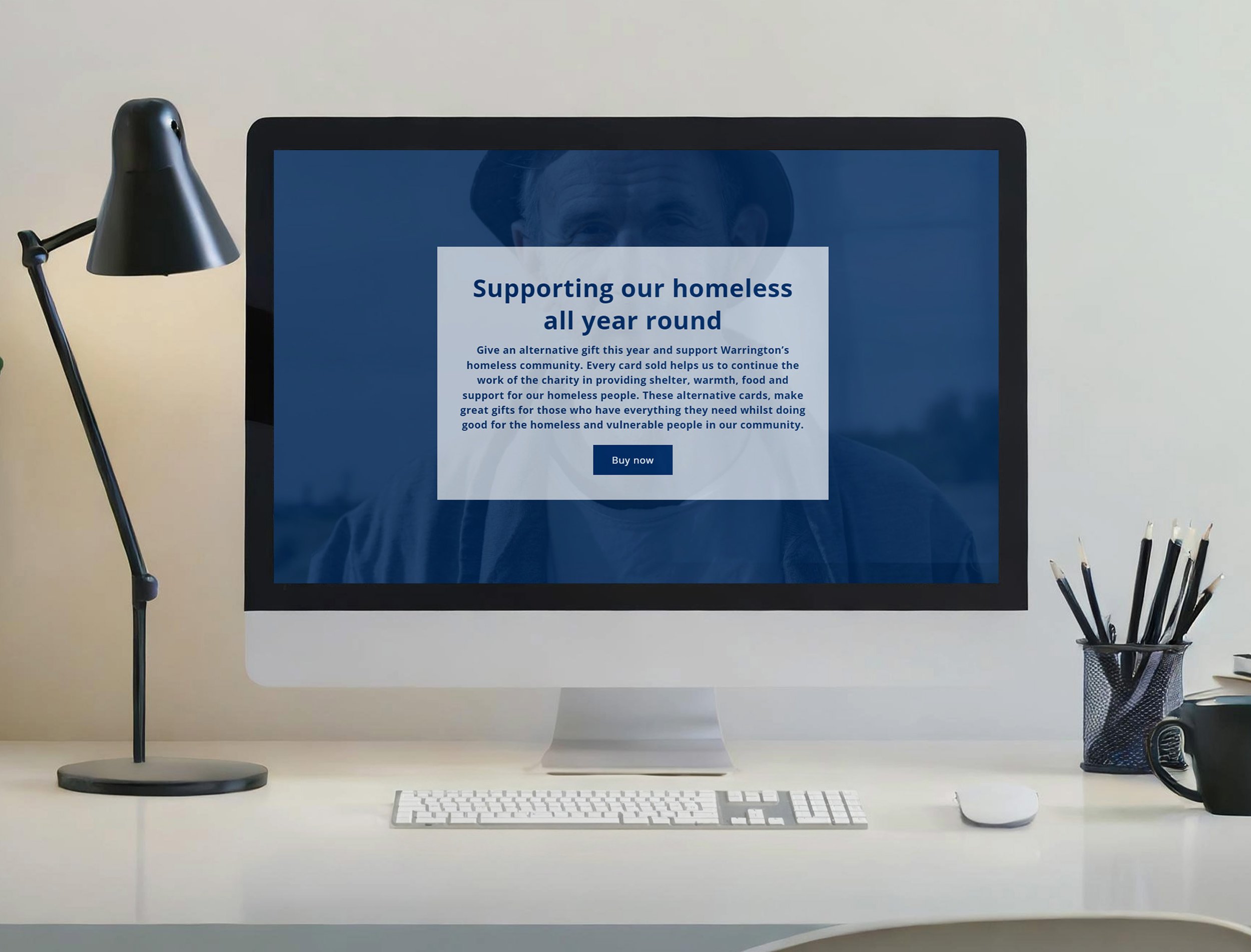 A computer monitor on a desk displaying a webpage supporting homeless charities, with a keyboard, mouse, desk lamp, pen holder, and mug on the desk.