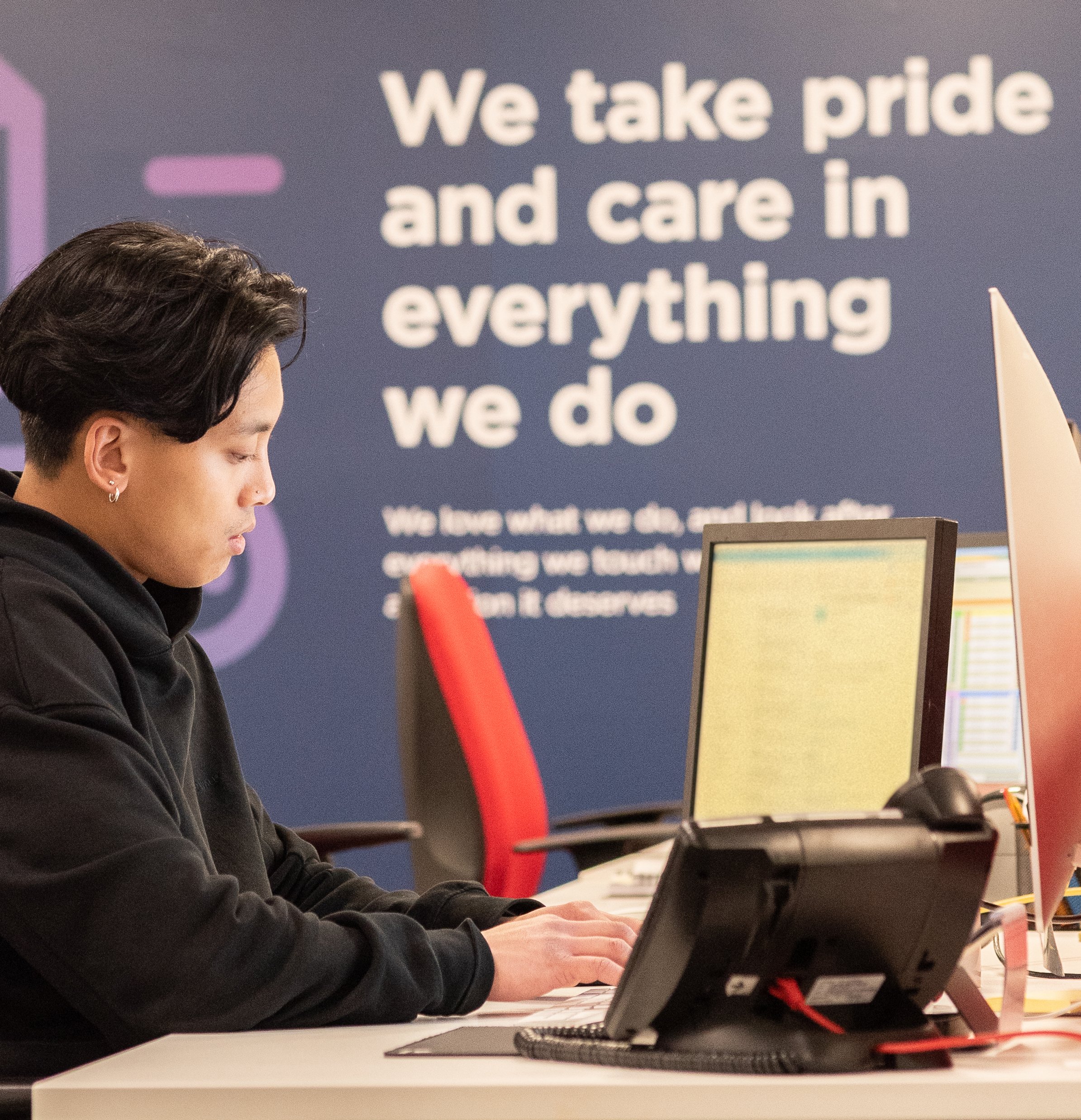 A person with short dark hair and earrings sitting at a desk working on a computer, with a large sign behind them that reads 'We take pride and care in everything we do'.