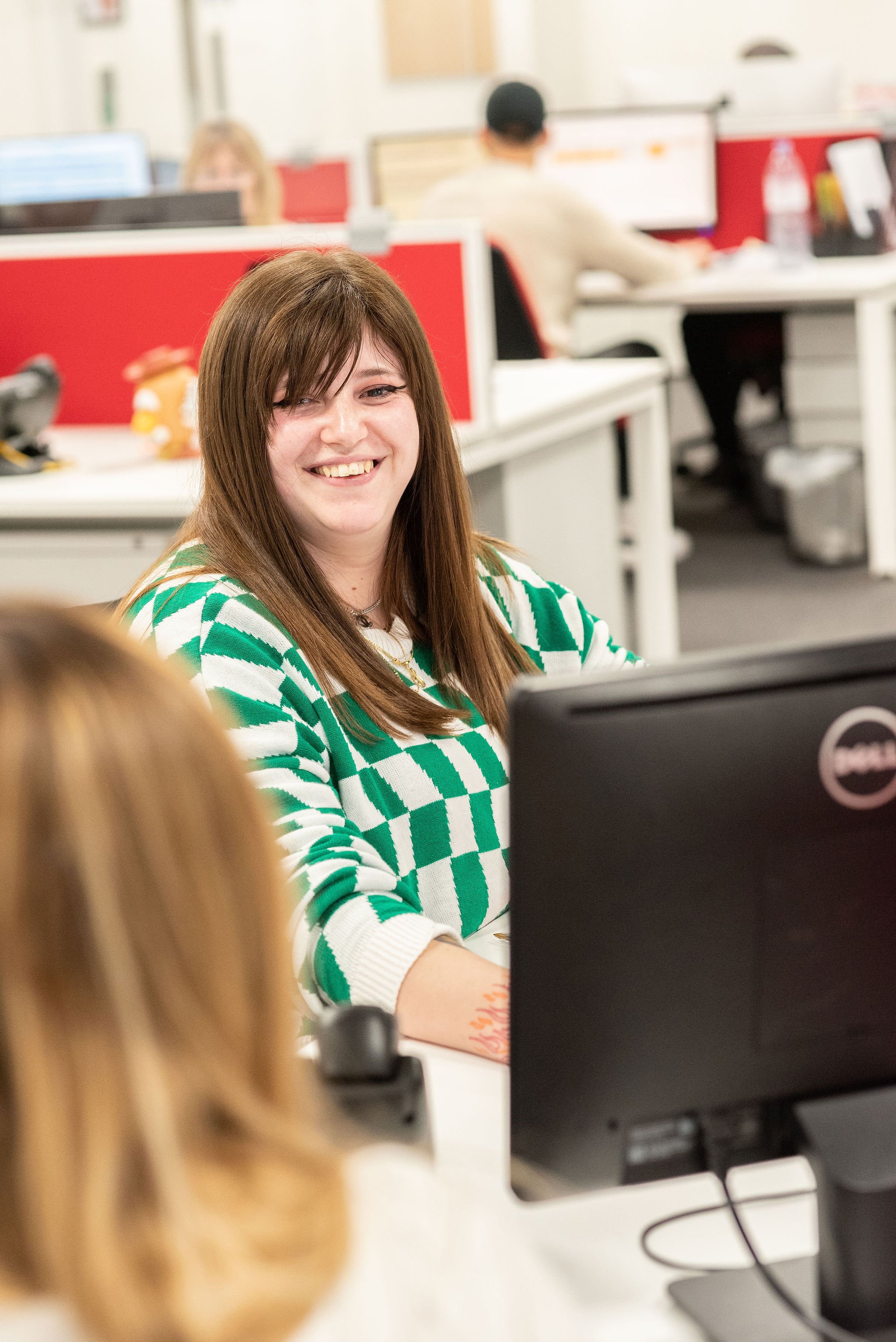Smiling woman with brown hair and green-and-white striped sweater sitting at a desk in an office with computer monitors, other employees, and a red cubicle wall in the background.