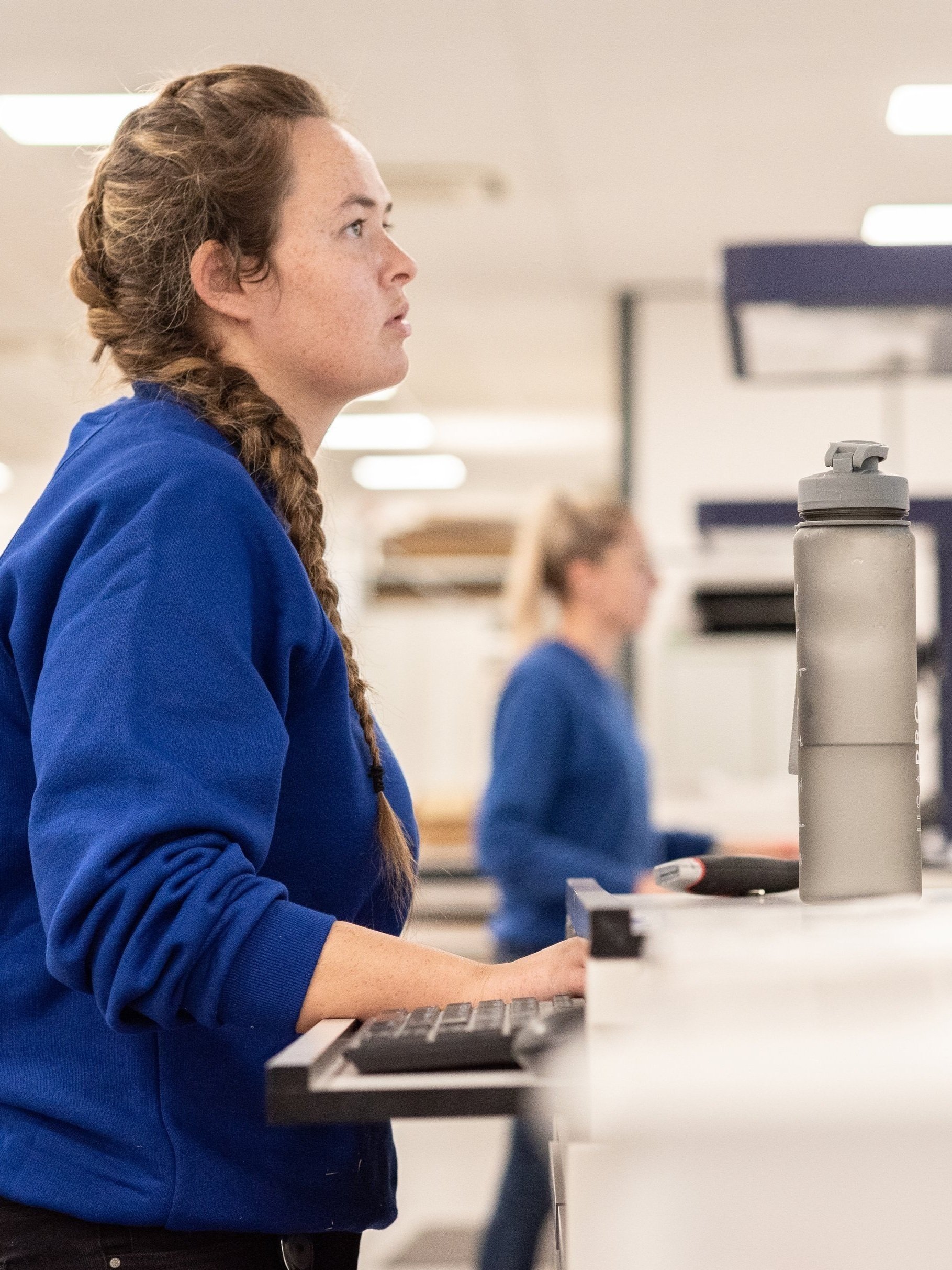 A woman with braided reddish-brown hair wearing a blue sweatshirt working at a checkout counter with a water bottle and cash register in front of her, in a retail store.