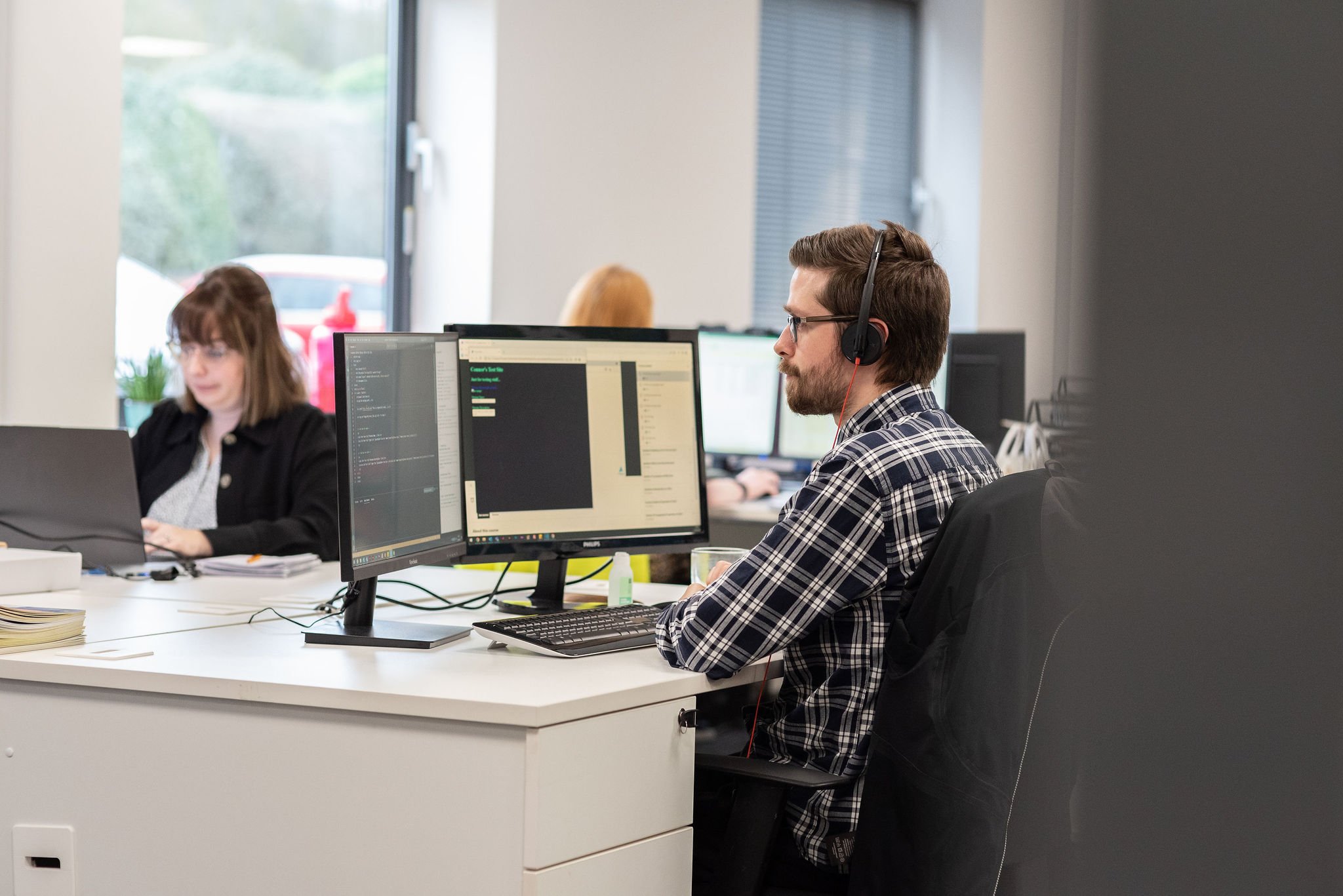 A man with glasses, a beard, and wearing a headset, working on a computer in an office setting with multiple monitors; a woman working on a laptop and other people working at desks in the background.