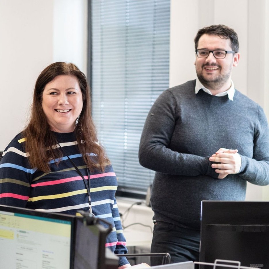 A woman with long brown hair wearing a striped sweater and a man with glasses and a beard wearing a gray sweater, standing in an office with computers and windows with blinds, smiling and engaging in conversation.