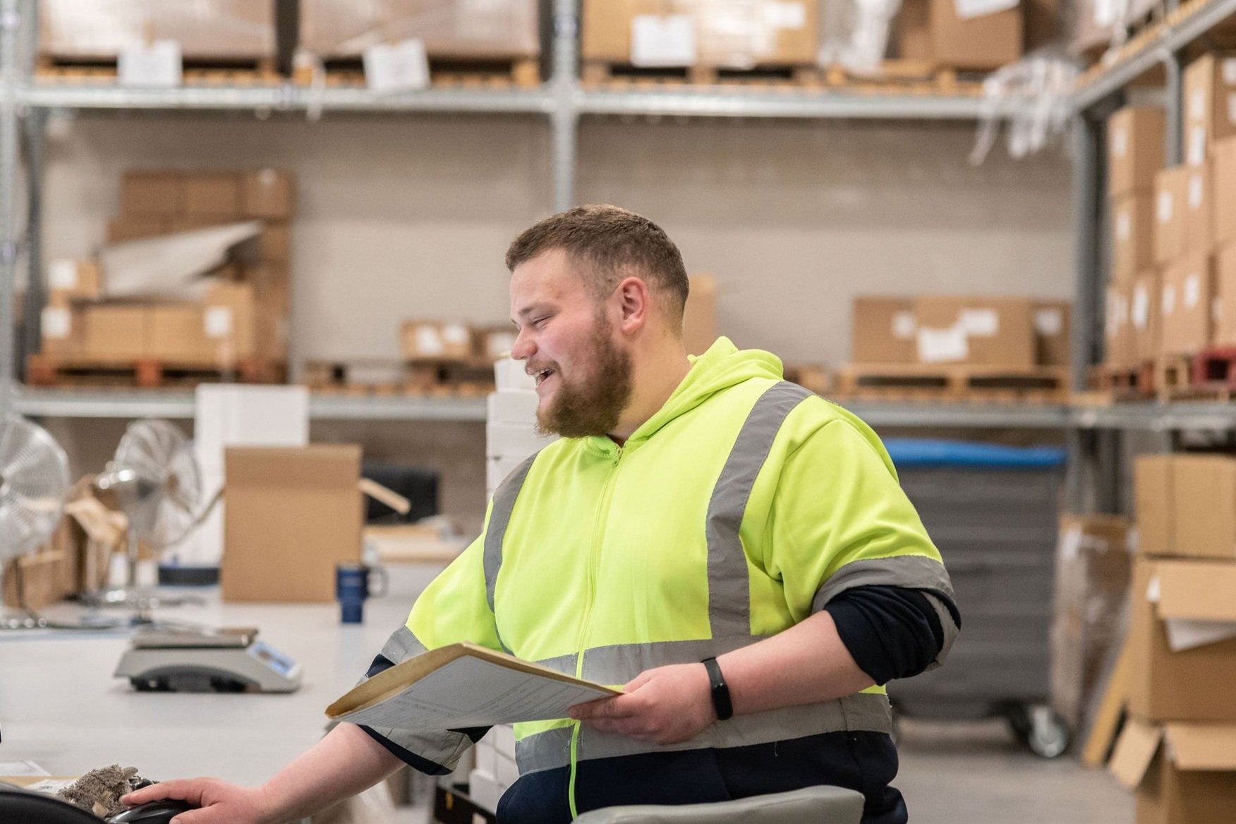 A man in a high-visibility yellow safety jacket sitting at a desk in a warehouse, smiling and holding a clipboard.