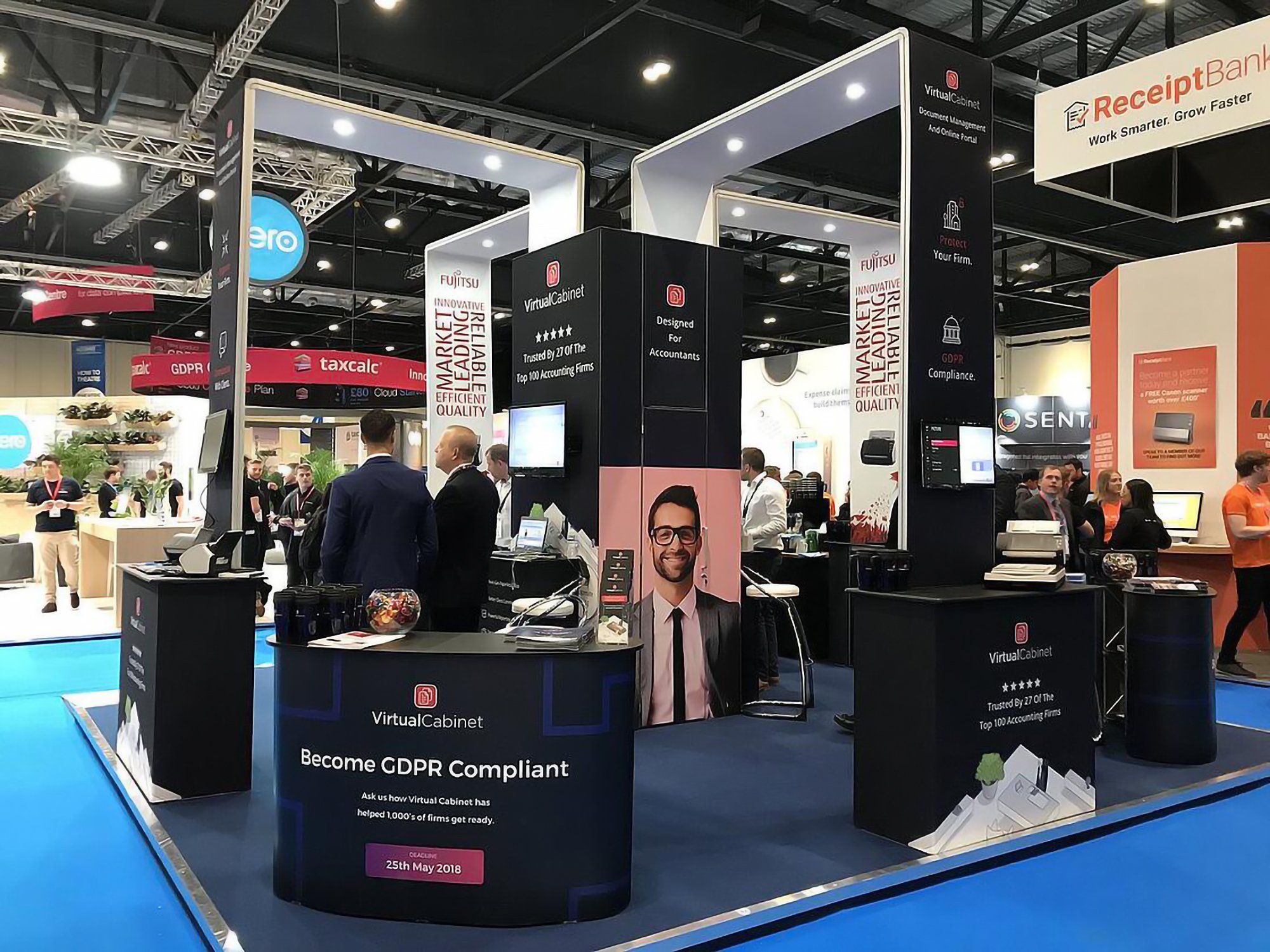 Trade show booth for VirtualCabinet with banners and display screens, promoting GDPR compliance and accounting firm services, set in a large exhibition hall with other booths in the background.