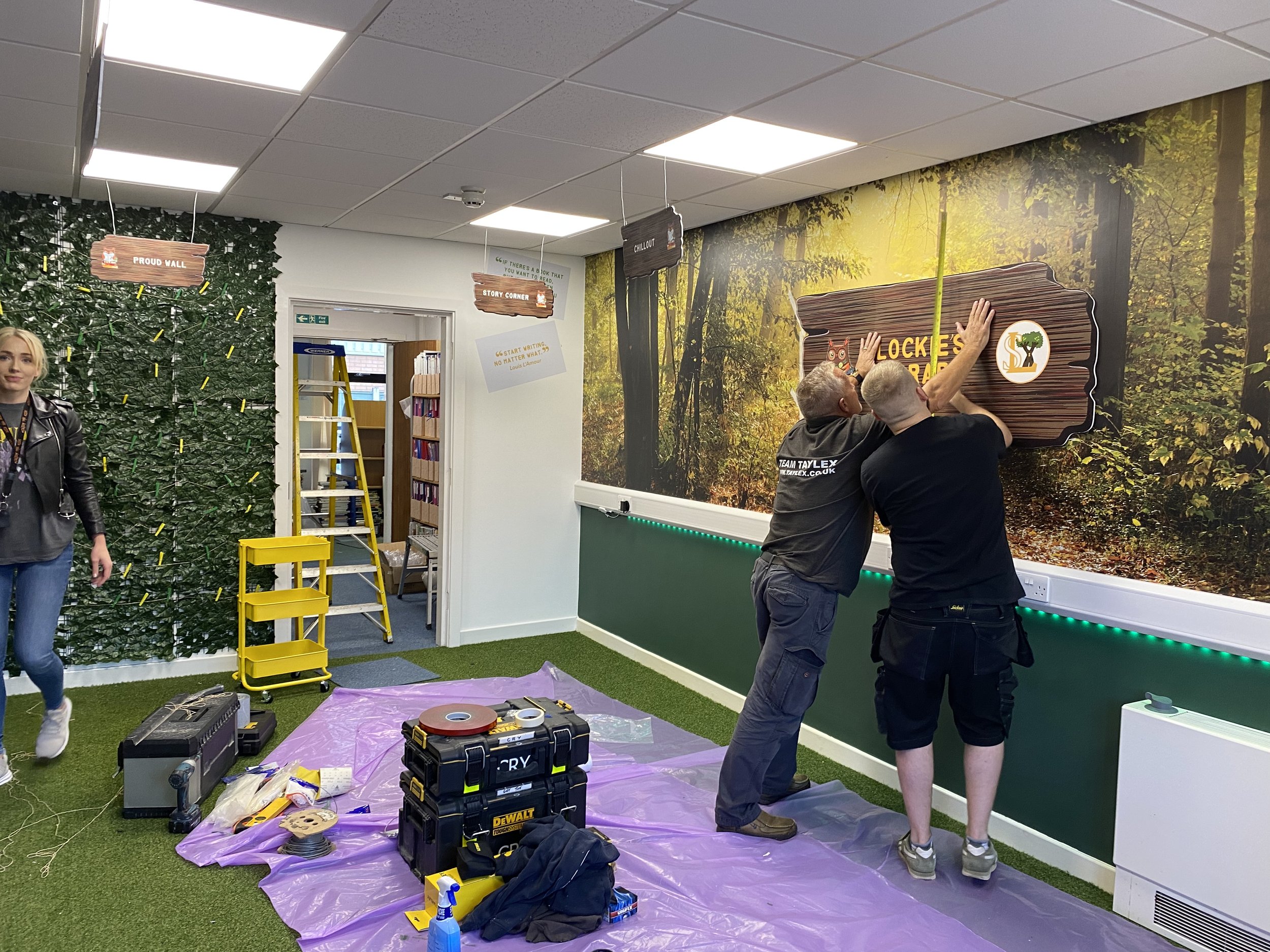 Two men installing a sign that says "Lockers" on a forest-themed wall in an indoor room. A woman walks past on the left, and there are tools and equipment on a purple tarp on the floor.
