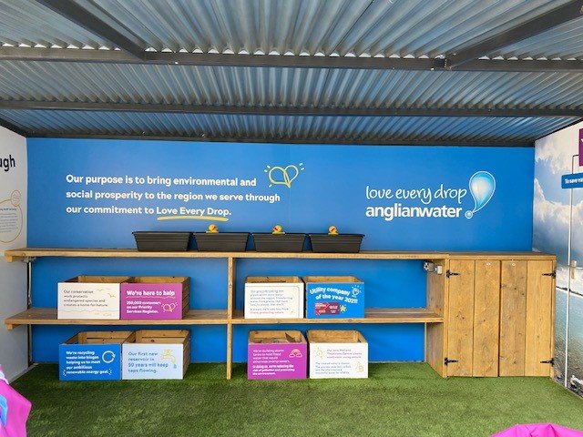 Blue informational booth with plants in pots on a shelf, and boxes with informational signs about environmental initiatives, under a corrugated metal roof.