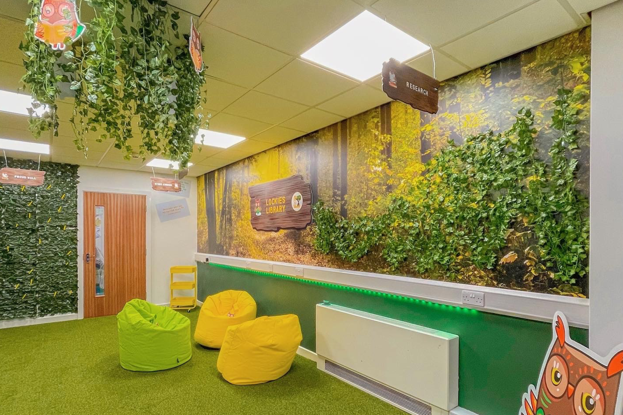 A colorful indoor play area with bean bags, green and yellow in color, and a nature-themed wall mural featuring trees and foliage. There are hanging decorations and signs, including one that says 'Research.' The room has a grassy green carpet and overhead lighting.