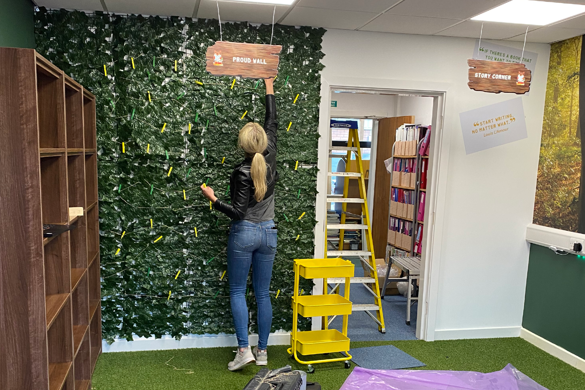 A woman standing on a step ladder, placing a sign that reads 'PROUD WALL' on a green leafy wall with clothespins. Nearby are bookshelves and a yellow rolling cart, with toolbox and purple tarp on the green carpeted floor.